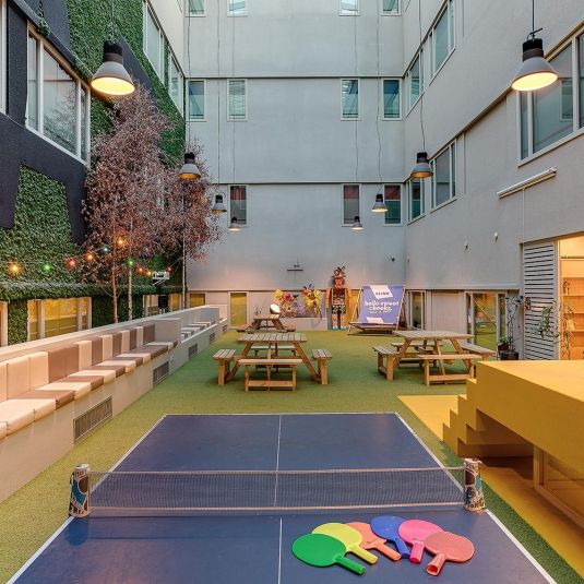 The Atrium at ClinkNOORD Hostel in Amsterdam, filled with a table tennis table, picnic benches, cushioned seating, plants and colour.